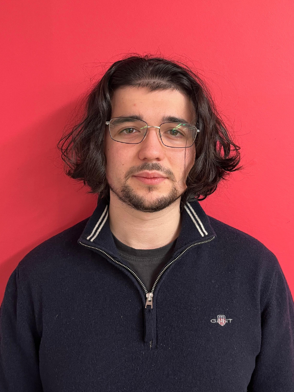 Your Marketing Team's Miguel Antonio is pictured standing in front of a pink wall. He has long brown hair and is wearing glasses. He is wearing a quarter zip and smiling.