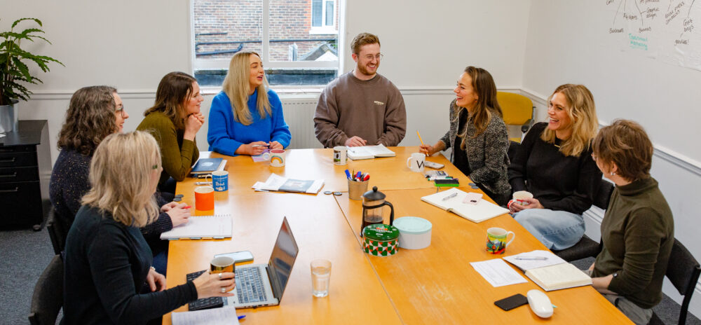 Your Marketing team are sat around a table in a meeting. The team are laughing.