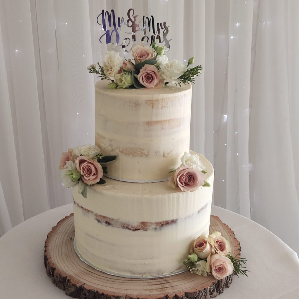 Two-tier white wedding cake with pink and white flowers. The cake is on a wooden base. There is an Mr. & Mrs cake topper on the top of the cake. 