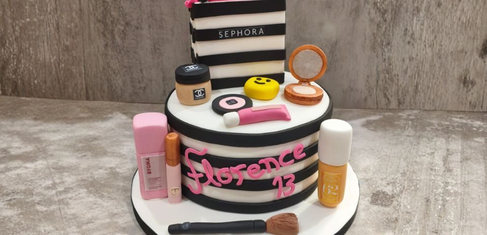 Sephora branded two tier cake. The cake has black and white stripes with a classic Sephora bag on the top of the cake. The cake is accessorised with a Chanel powder, Sephora makeup brush, Charlotte Tilbury mascara and Sol de Janeiro body spray.