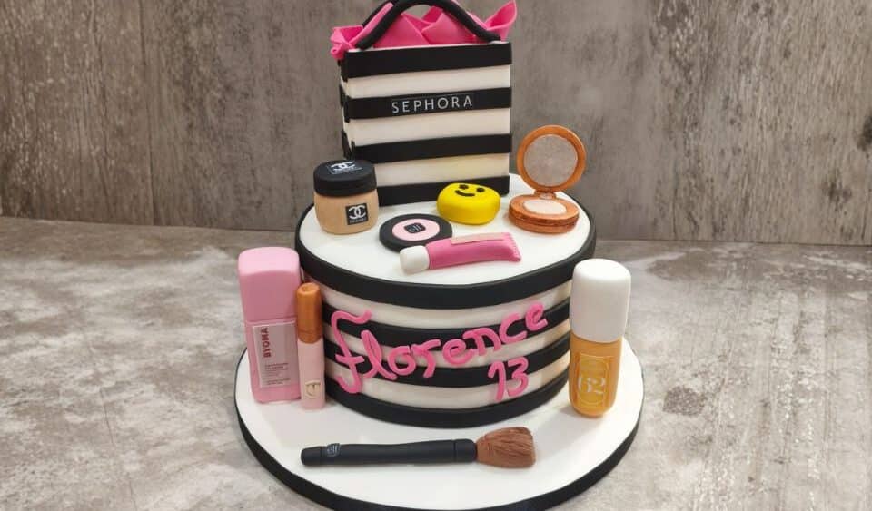 Sephora branded two tier cake. The cake has black and white stripes with a classic Sephora bag on the top of the cake. The cake is accessorised with a Chanel powder, Sephora makeup brush, Charlotte Tilbury mascara and Sol de Janeiro body spray.
