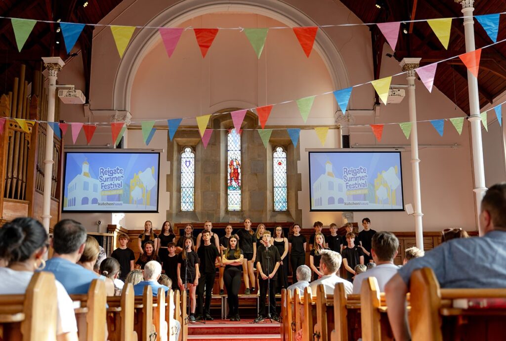 A choir is pictured singing inside a church in Reigate at Reigate Summer Festival. There is multicoloured bunting hung across the church. 