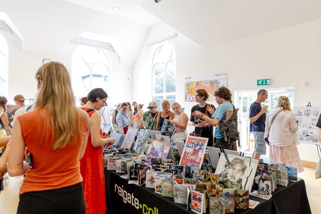 People are pictured browsing art created by Reigate College. There is a table with a black Reigate College-branded tablecloth with lots of canvas' on top. 