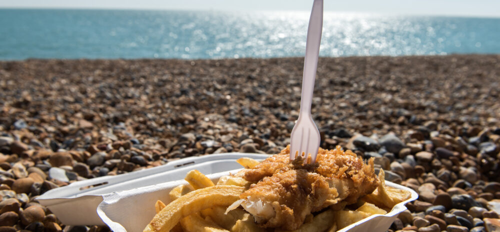 A white container filled with traditional british fish and chips with a fork resting on top. The sea, blue sky and pebbles are pictured in the background. User search behaviour at BrightonSEO 2026.