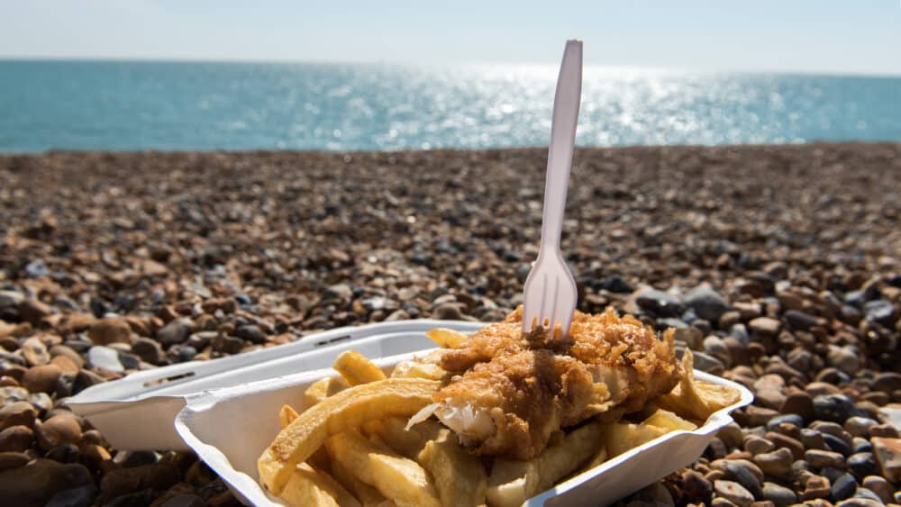 A white container filled with traditional british fish and chips with a fork resting on top. The sea, blue sky and pebbles are pictured in the background. User search behaviour at BrightonSEO 2026.