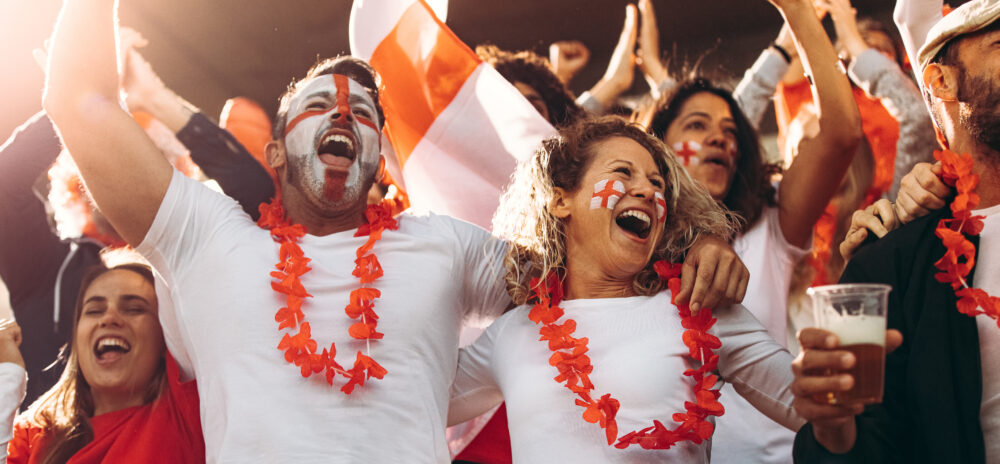 English fans cheering the World Cup football matches in white tees with St George crosses painted on their faces, waving flags.