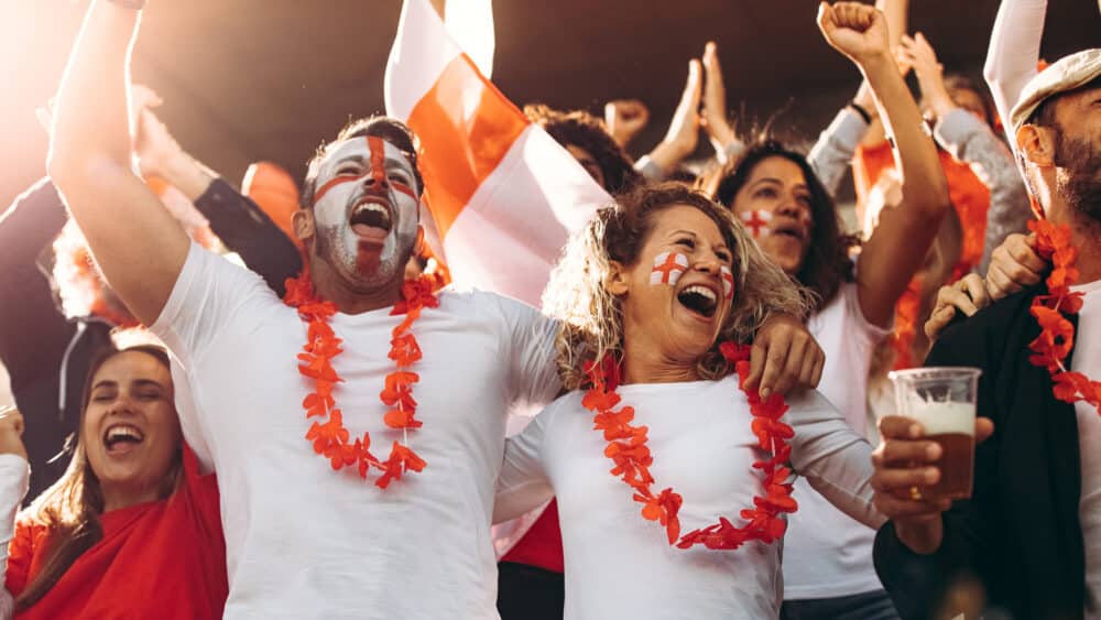 English fans cheering the World Cup football matches in white tees with St George crosses painted on their faces, waving flags.