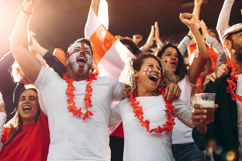 English fans cheering the World Cup football matches in white tees with St George crosses painted on their faces, waving flags.