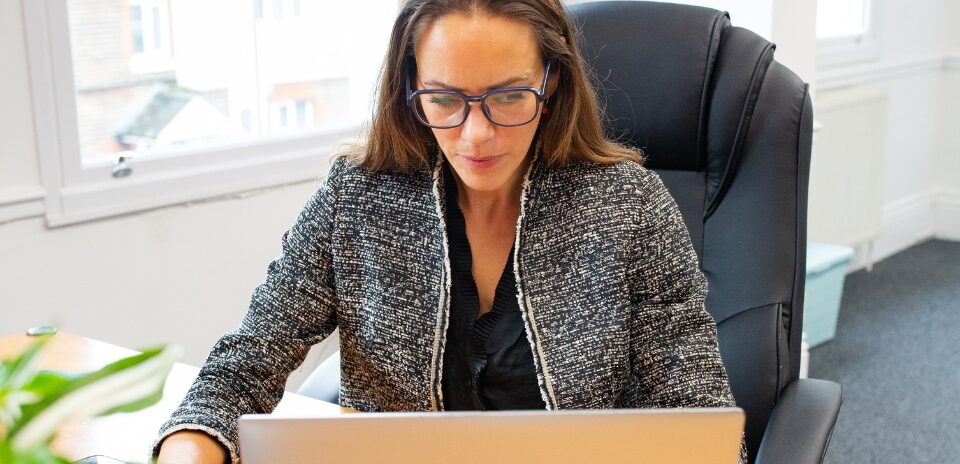Laura Taylor is sat at her desk looking at her laptop, wearing a grey jacket, black top.